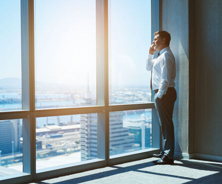 Confident businessman looking out of large windows at a view of the city below while talking on his mobile phone
