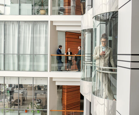 Busy people working in business hall. Three colleagues discussing project, young woman checking phone in glassy elevator