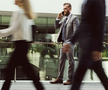 A bearded man in a gray suit stands in a modern glass-lined building, talking on a phone as blurred people walk past in the foreground