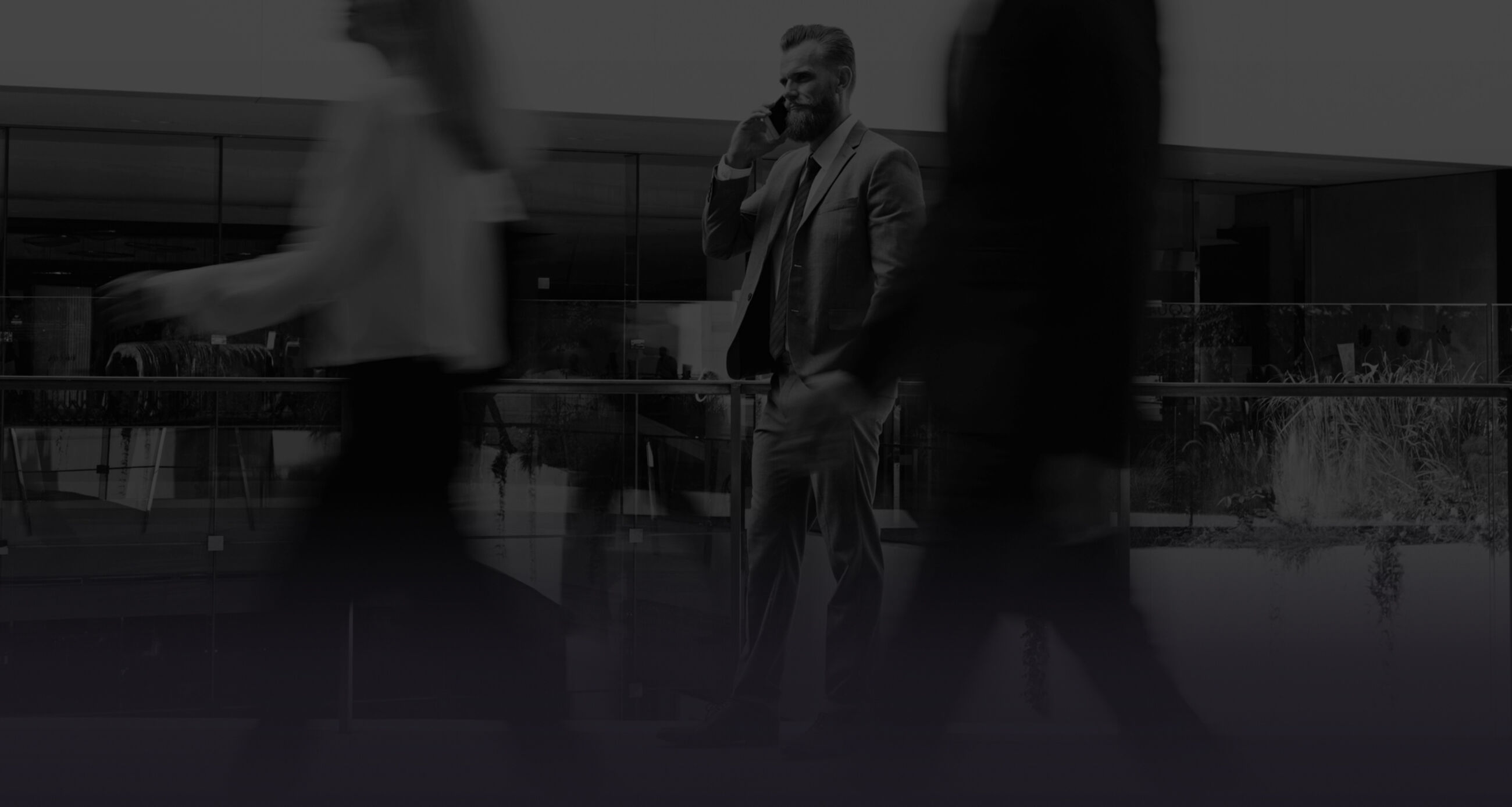 A bearded man in a gray suit stands in a modern glass-lined building, talking on a phone as blurred people walk past in the foreground